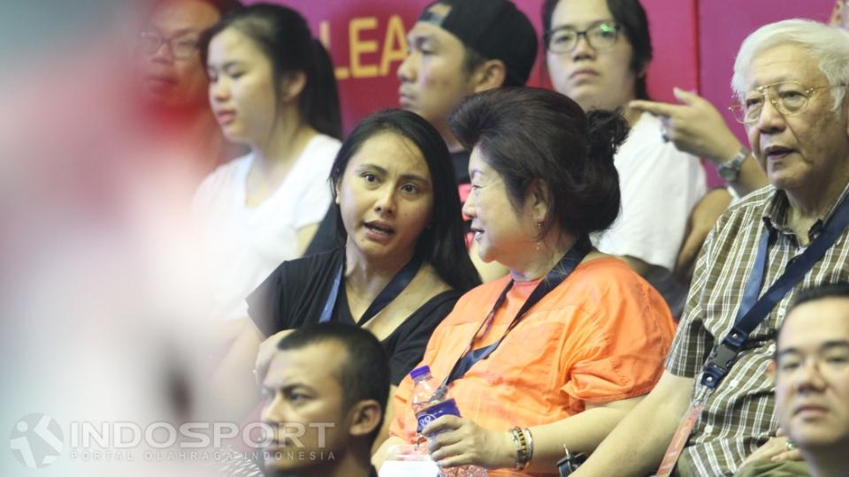 Wanita berbaju hitam menyaksikan pertandingan sambil berbincang dengan orang yang di sebelahnya.