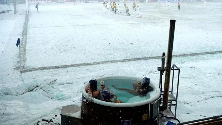 Meski di tengah cuaca salju, tiga wanita menonton laga Hobro vs Copenhagen dari dalam kolam air panas di pinggir lapangan. Copyright: Getty Images.