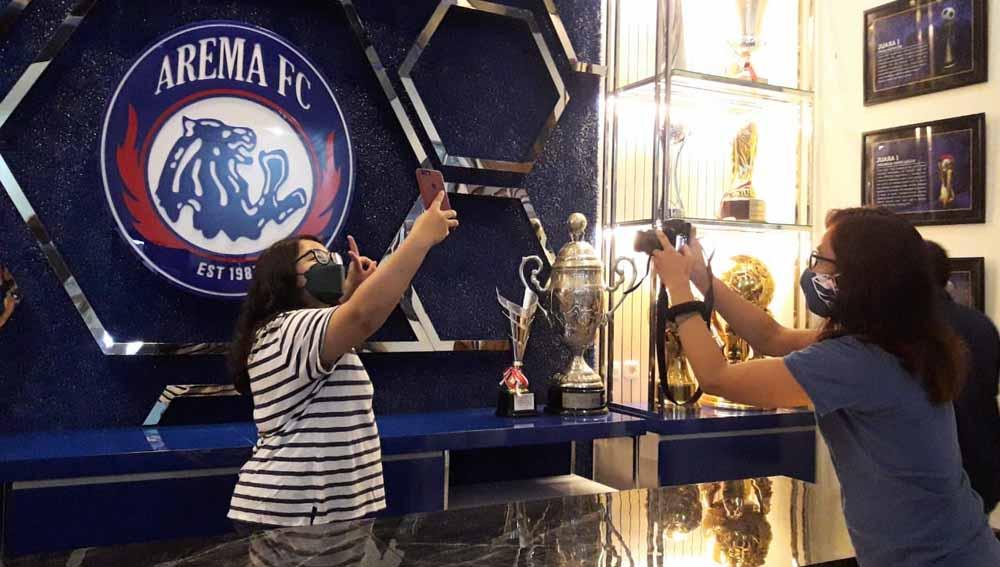 Pengunjung Arema Champions Corner berfoto dgn latar belakang logo dan deretan piala.