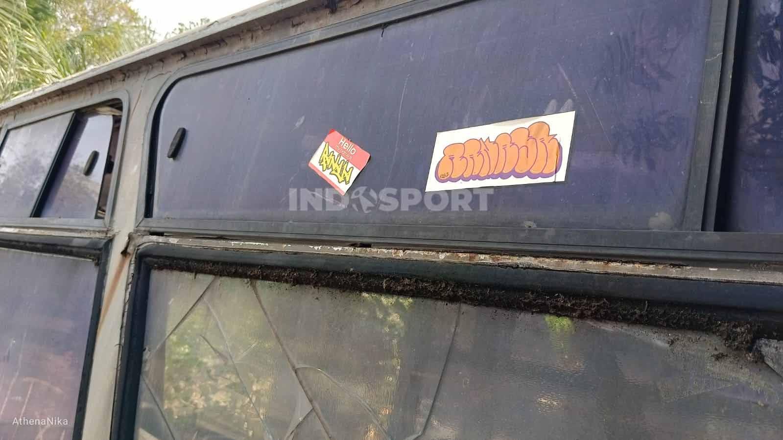 Stiker yang menempel di kaca bus tampak masih baru hasil 'karya' anak muda sekitar. Beberapa kaca samping bahkan sudah pecah. (Foto: Indra Citra Sena/INDOSPORT)