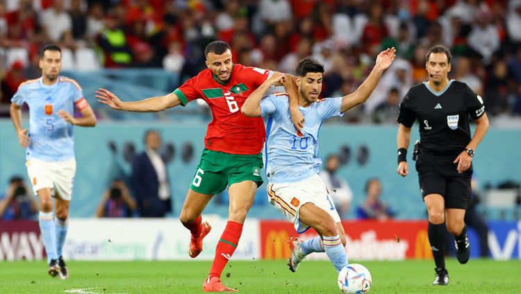 Duel fisik terjadi antara striker Spanyol, Marco Asensio (kanan) dengan bek Maroko, Roman Saiss dalam pertandingan babak 16 besar Piala Dunia 2022 (Foto: REUTERS/Bernadett Szabo). Copyright: © REUTERS/Bernadett Szabo